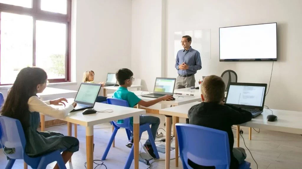 Teacher standing and explaining in front of children using laptops in a modern classroom.