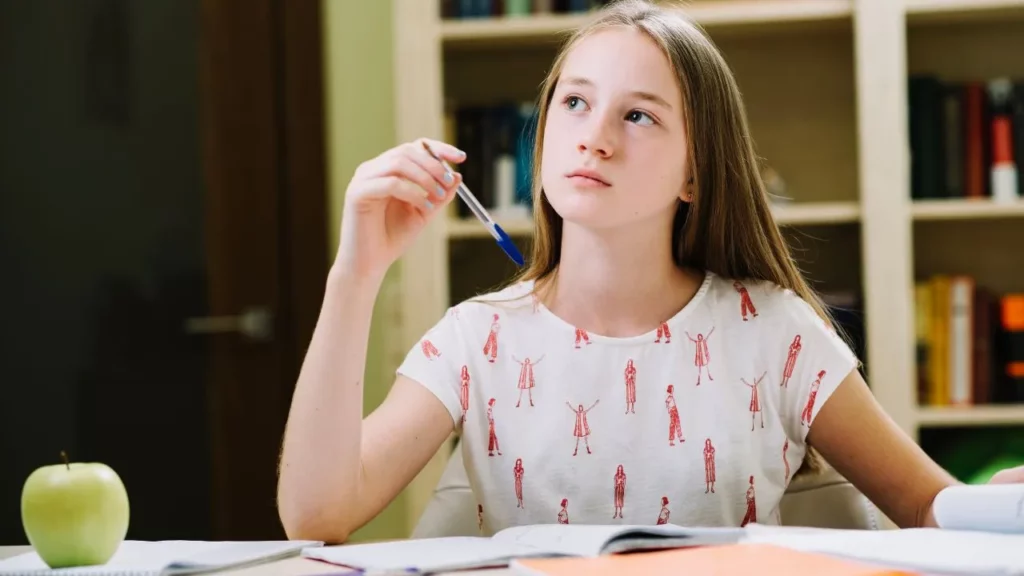 Thoughtful young girl at desk holding a pen, looking up while studying with books and an apple.