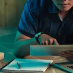 Person in a blue shirt lying on the floor, focused on a glowing tablet surrounded by open books and stationery.