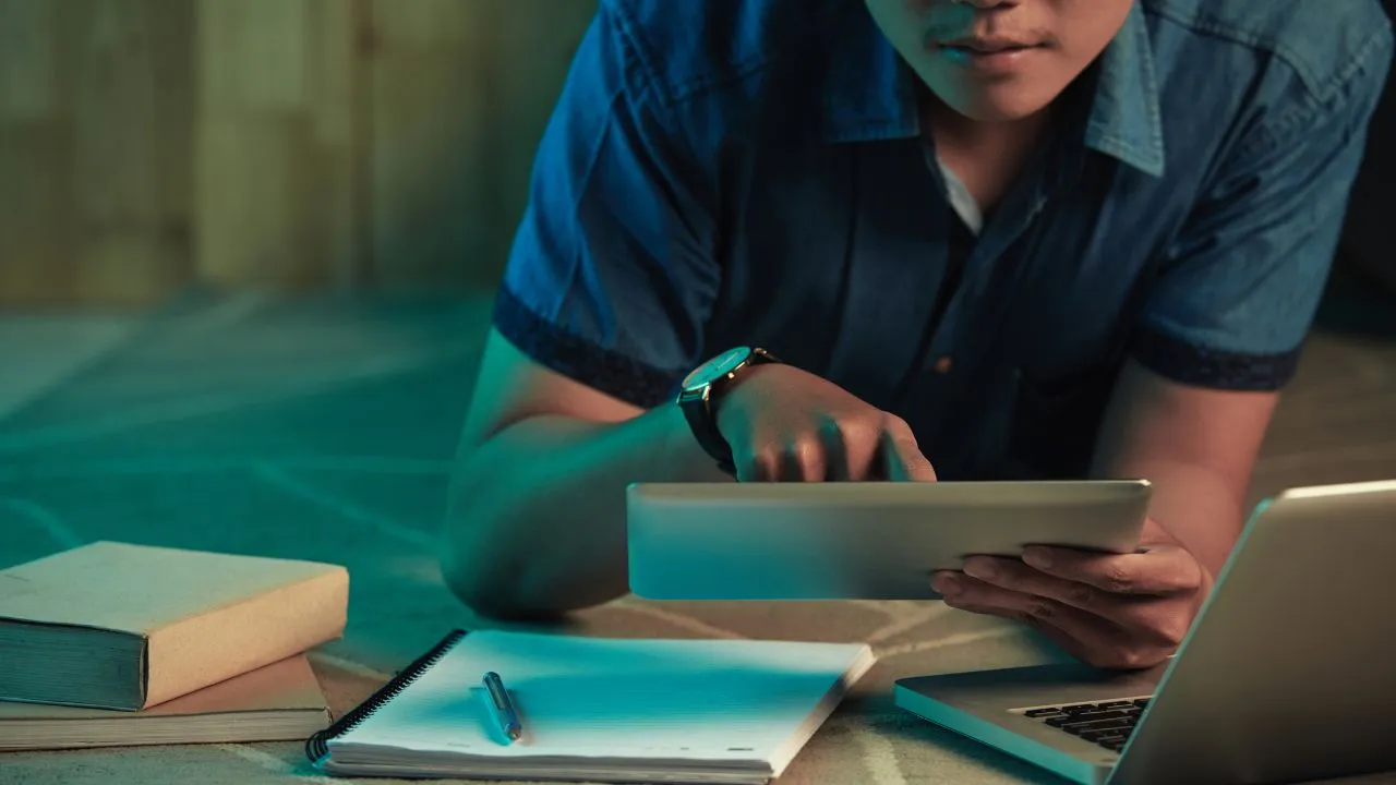 Person in a blue shirt lying on the floor, focused on a glowing tablet surrounded by open books and stationery.