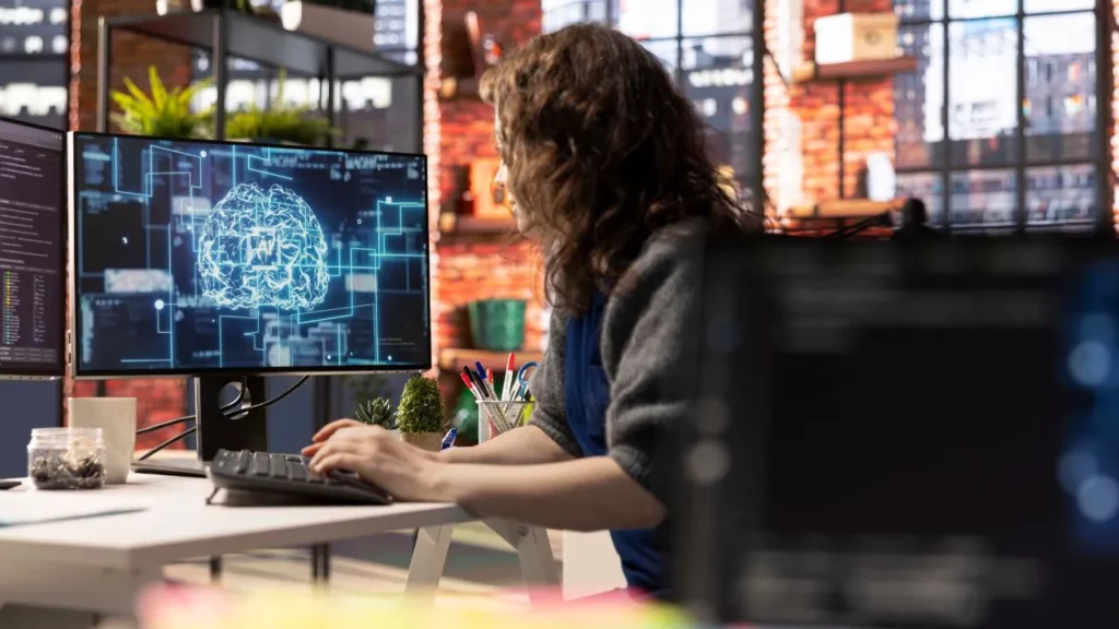 Woman in a modern office working intently at a large monitor displaying an AI neural network visualization.