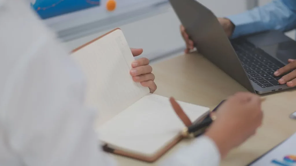 Close-up of hands taking notes in a small notebook during a business meeting, with a laptop open nearby.