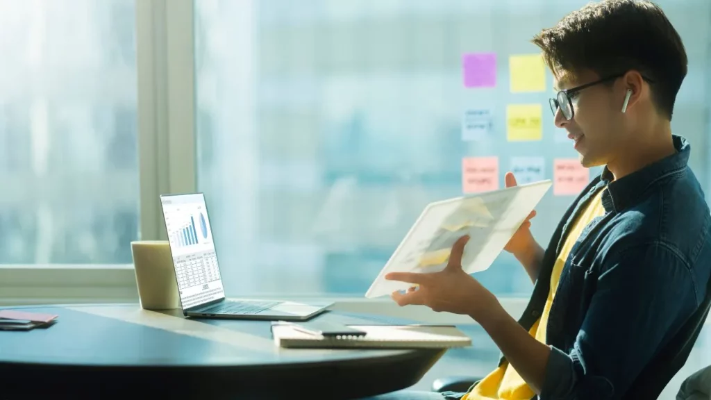 Young man in casual clothes smiling while reviewing printed charts and using a laptop at a desk with sticky notes on the wall.