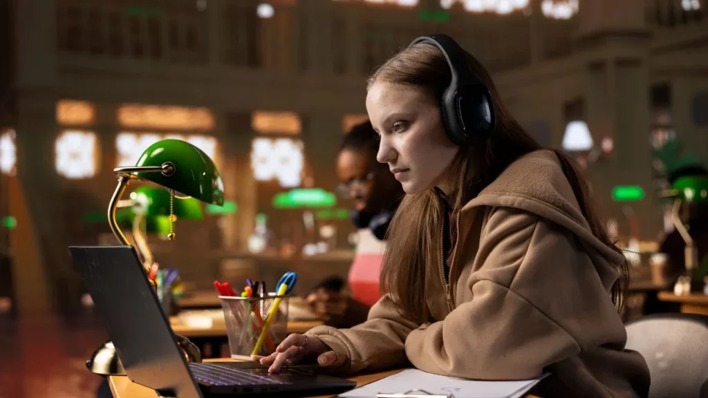 Teenage girl wearing large over-ear headphones, intently working on a laptop in a warmly lit library or study room with green desk lamps.