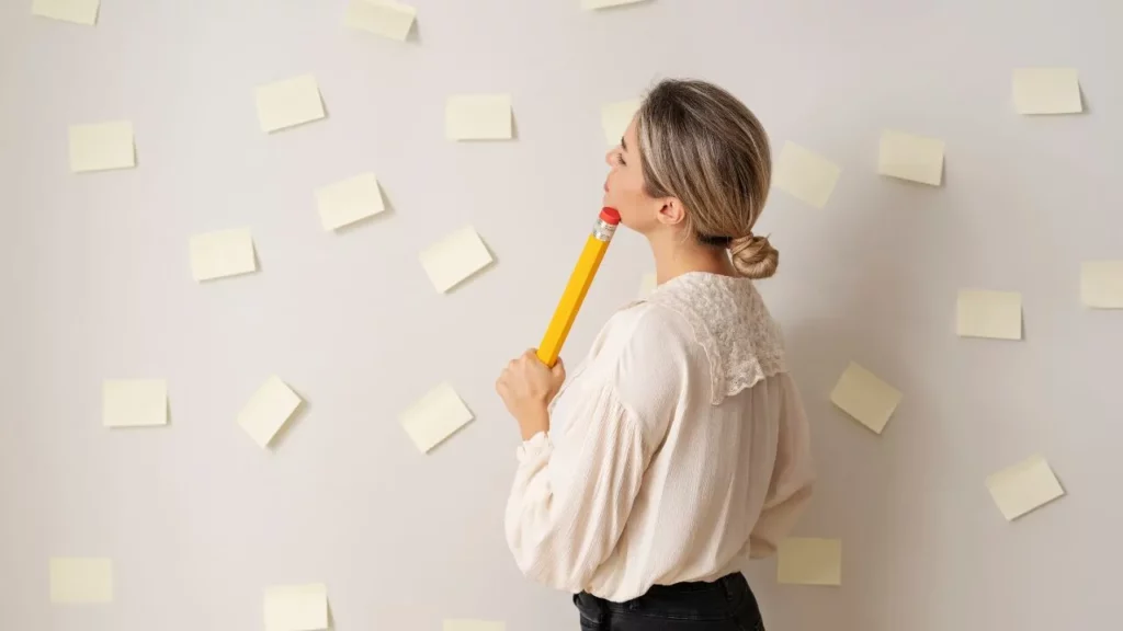 Woman in a white blouse holding a large yellow pencil in her mouth, thoughtfully looking up at a wall covered in sticky notes.