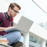 Young man with headphones and maroon sweater, working on laptop while sitting on campus steps.