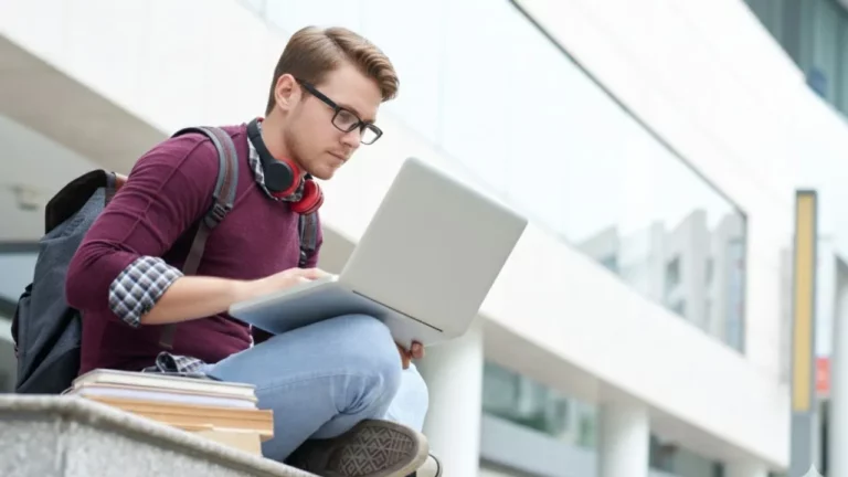 Young man with headphones and maroon sweater, working on laptop while sitting on campus steps.