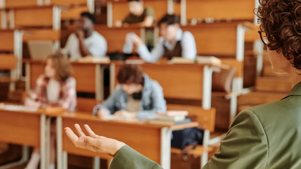 A lecturer in green blazer gesturing during university lecture to seated students.