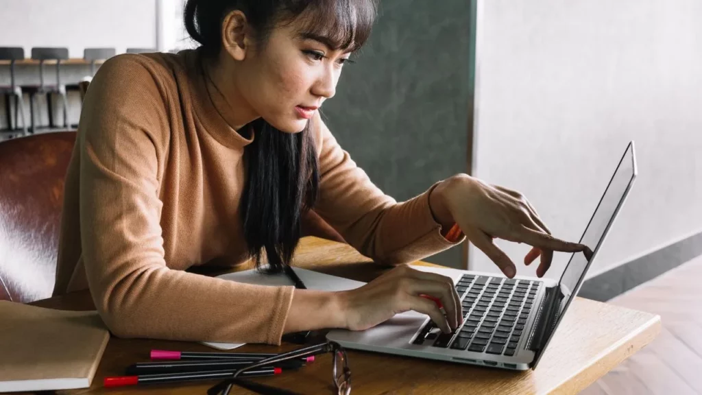 Young Asian woman in brown sweater, focused on her laptop in a study space.