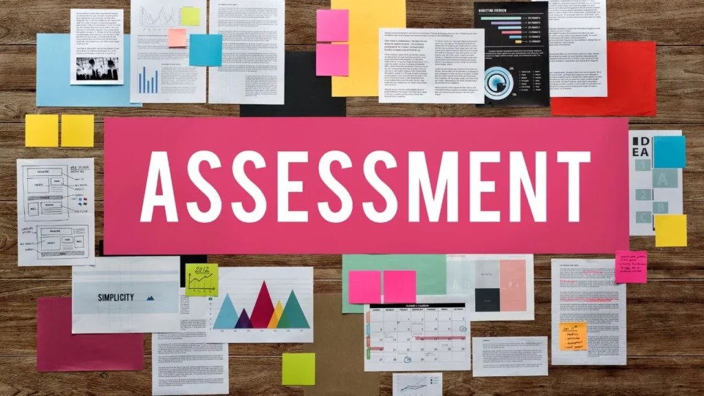 A wooden desk covered in various charts, notes, and a large pink sign that says "ASSESSMENT."