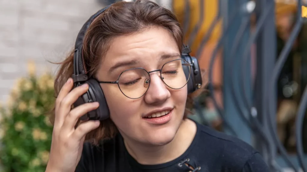 A person wearing large over-ear headphones and glasses, looking downward with a slight smile.