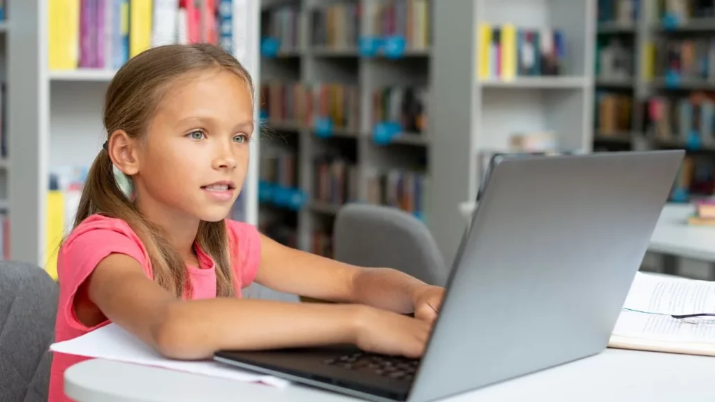 A young girl sitting in a library and focused on a laptop screen.