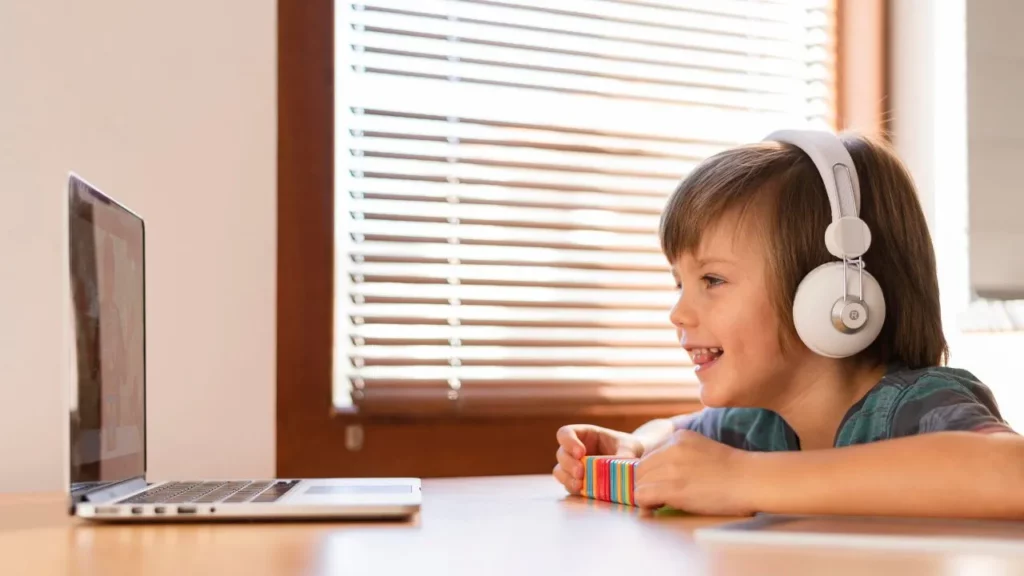 A smiling young boy wearing white headphones while watching a laptop screen at a table.
