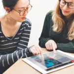 Two young women collaborating and reviewing global news on a digital tablet.