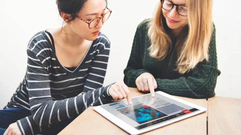 Two young women collaborating and reviewing global news on a digital tablet.