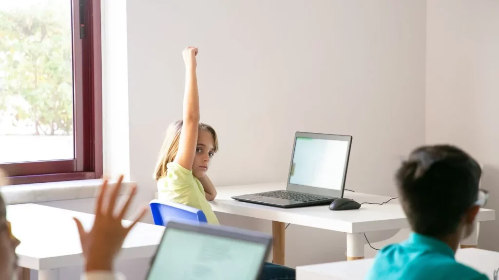 A young student raising her hand while working on a laptop in a classroom.
