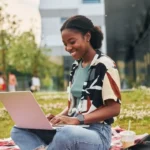 A student smiling while working on her laptop outdoors on a picnic blanket.