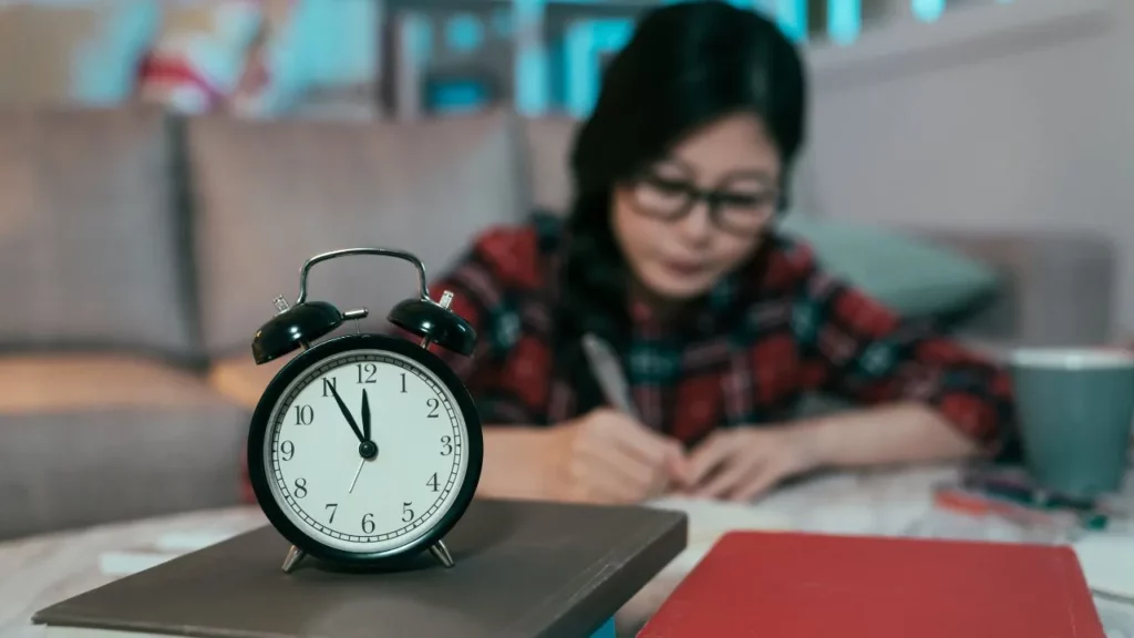 A student studying late into the night, with an alarm clock showing nearly 11:00.