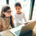 Two young girls looking at a laptop screen together by a window.