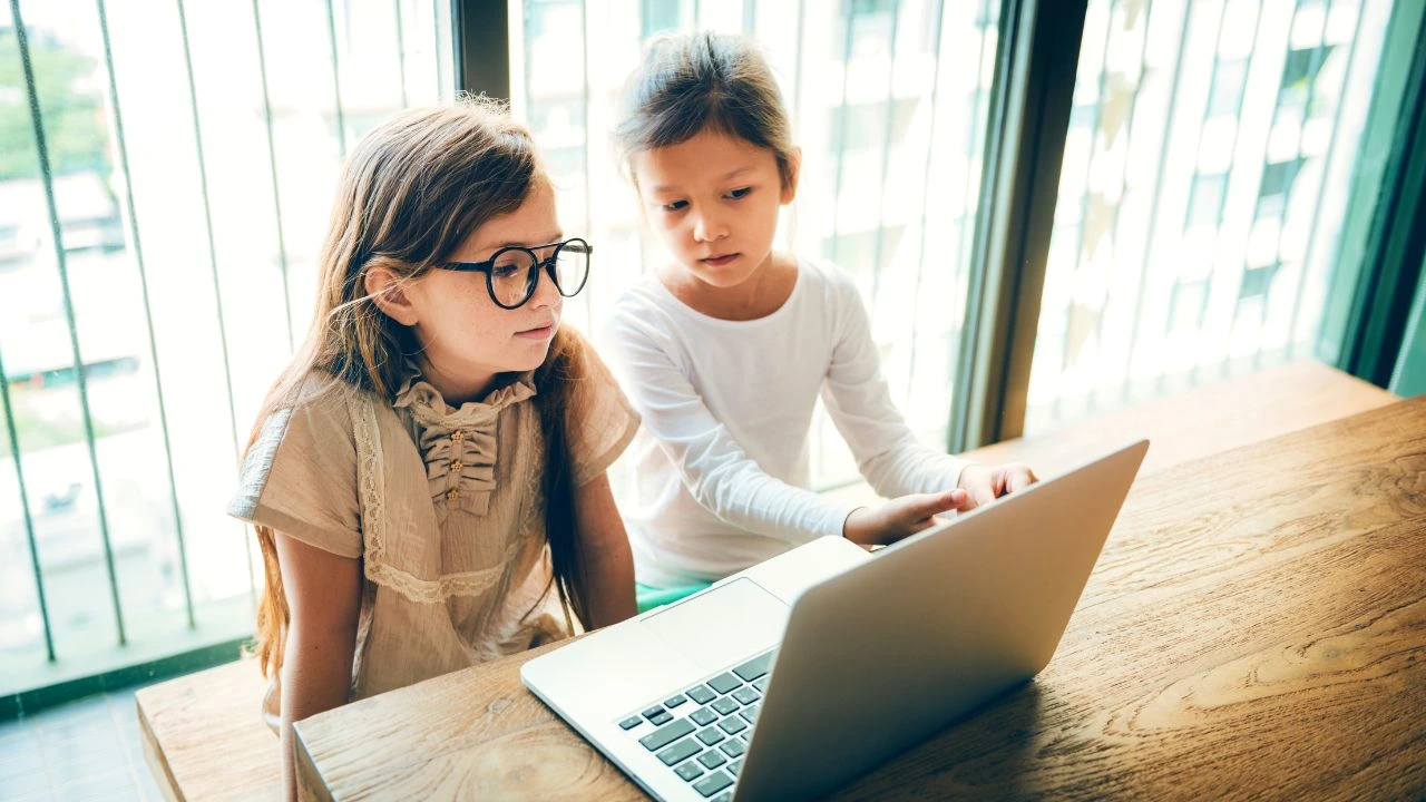Two young girls looking at a laptop screen together by a window.