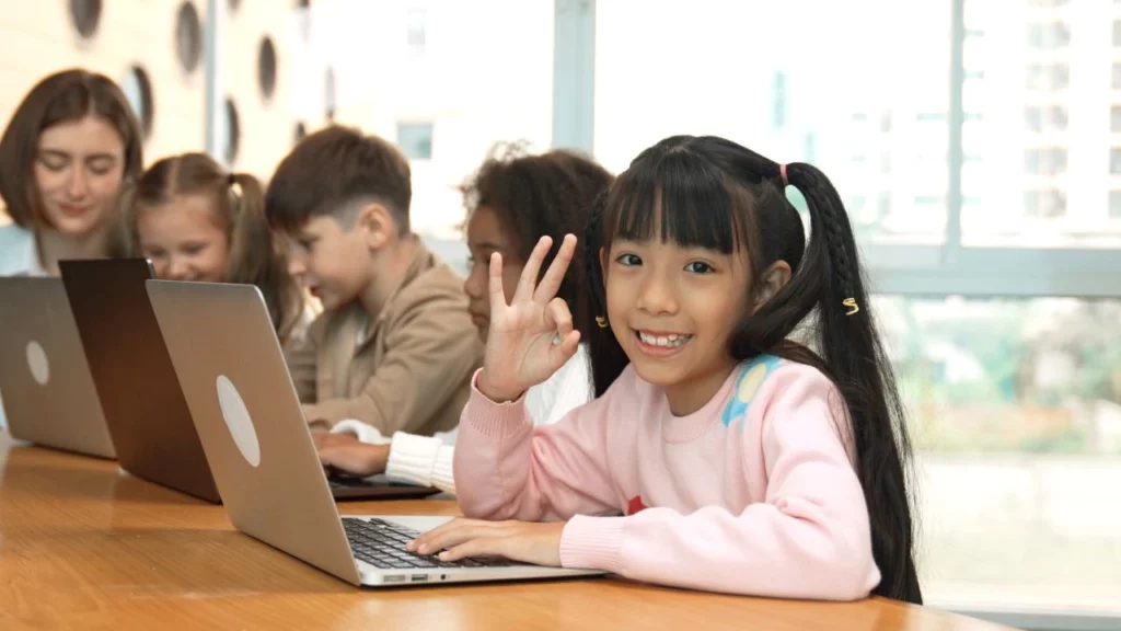 A smiling girl in a classroom giving an "OK" hand gesture while using a laptop.