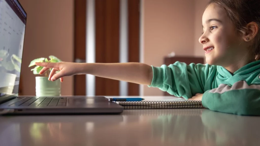 A young girl smiling and pointing at a laptop screen in a brightly lit room.
