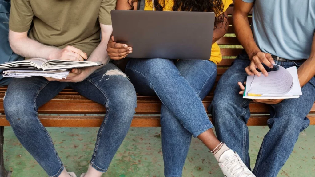 Three students sitting on a bench outdoors with a laptop and notebooks.