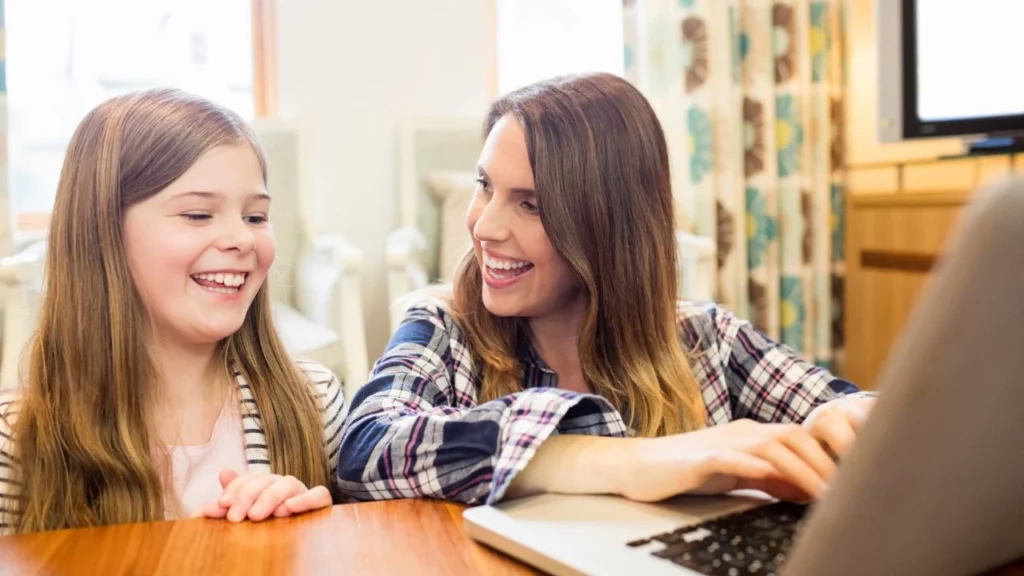 A woman and a young girl laughing while looking at a laptop together.