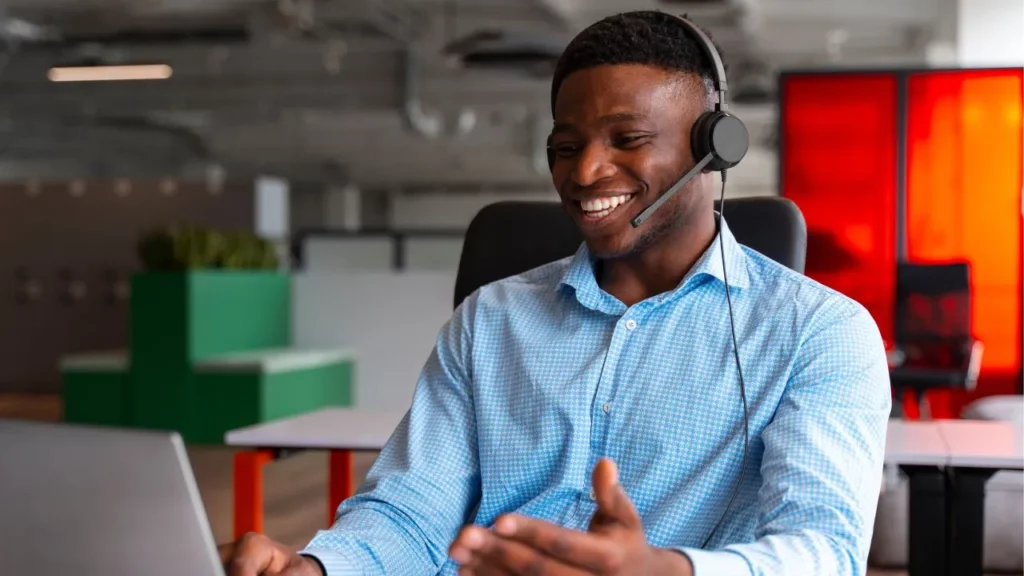 A smiling man wearing a headset and a blue patterned shirt gestures while participating in a video call on his laptop in a modern office.