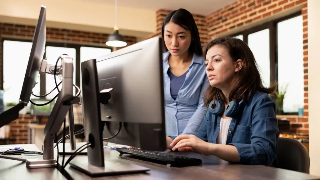 Two women in an office setting looking intently at a computer monitor while working together.