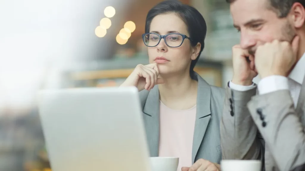 A woman with glasses and a man in a suit looking thoughtfully at a laptop screen.