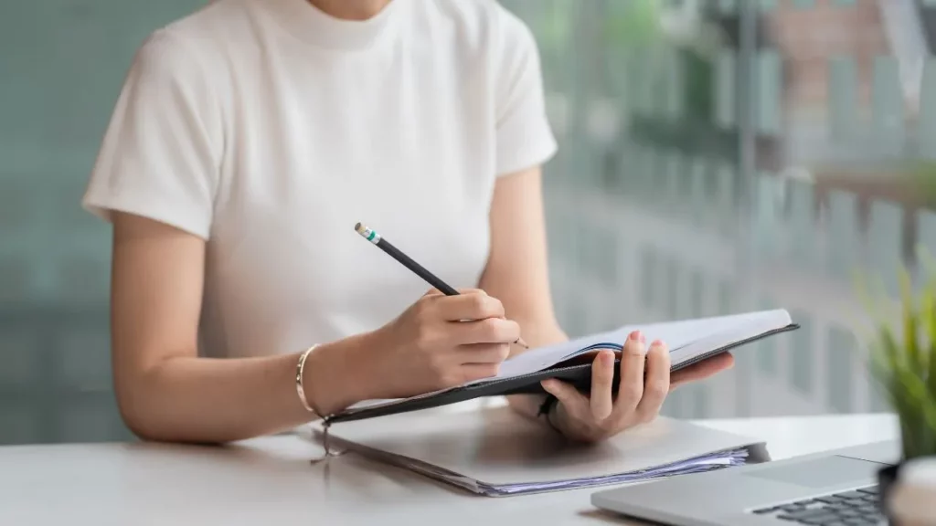Close-up of a person's hands writing with a pencil in a notebook at a desk.