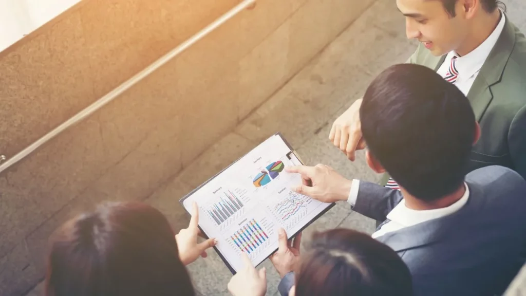 An overhead view of four professionals in business attire huddled together, pointing at and discussing various colorful charts and data visualizations on a clipboard.