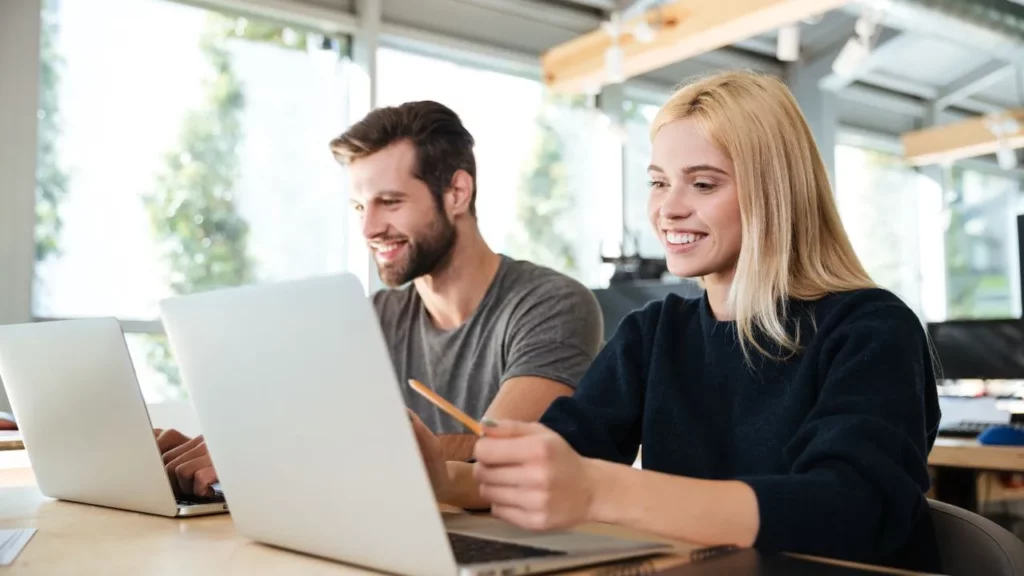 Two young professionals smiling while working on laptops in a bright, modern office space.