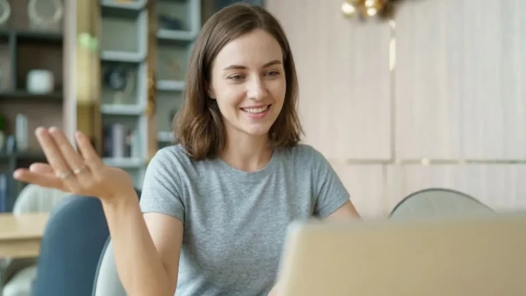 A smiling woman in a grey t-shirt sits in a brightly lit room, gesturing with her hand while looking at a laptop screen during a video call.