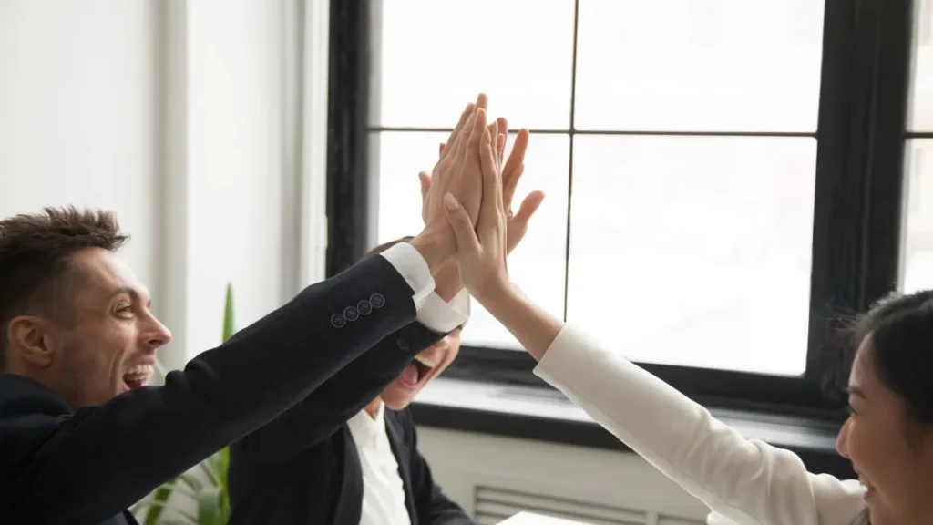 A diverse group of business professionals in suits giving a celebratory high-five in a bright office.