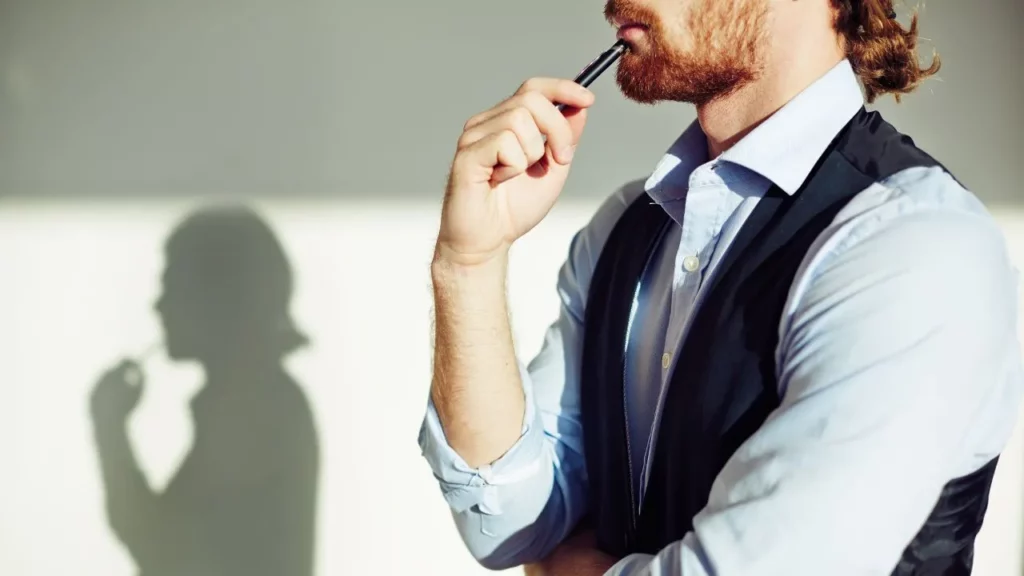 A close-up of a man with a beard and a vest holding a pen to his chin in a thoughtful, pensive pose.
