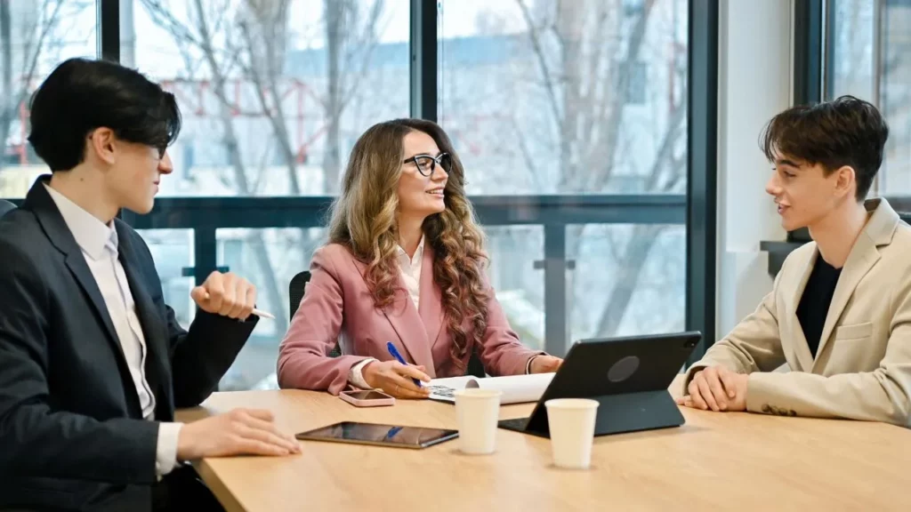 Three professionals in business attire having a meeting at a conference table with tablets and coffee.