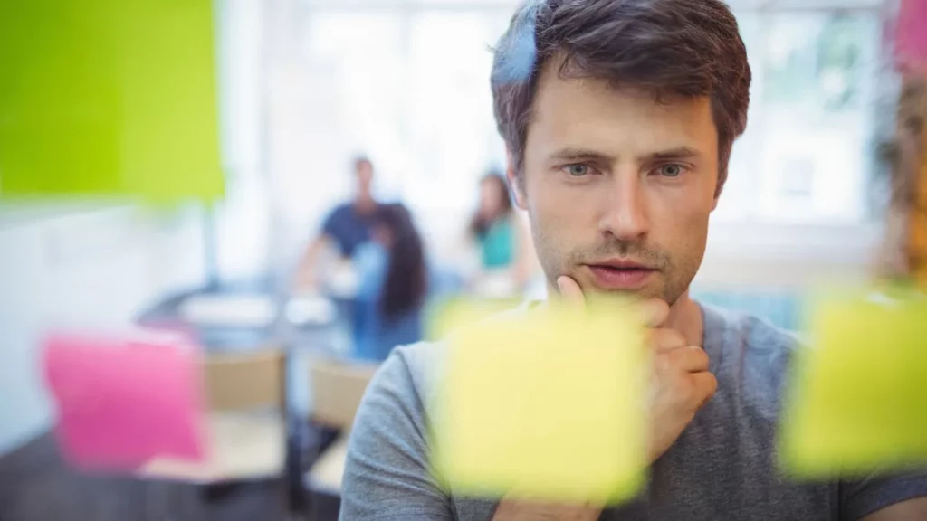 A man in a grey shirt looking thoughtfully at colorful sticky notes on a glass wall in an office setting.
