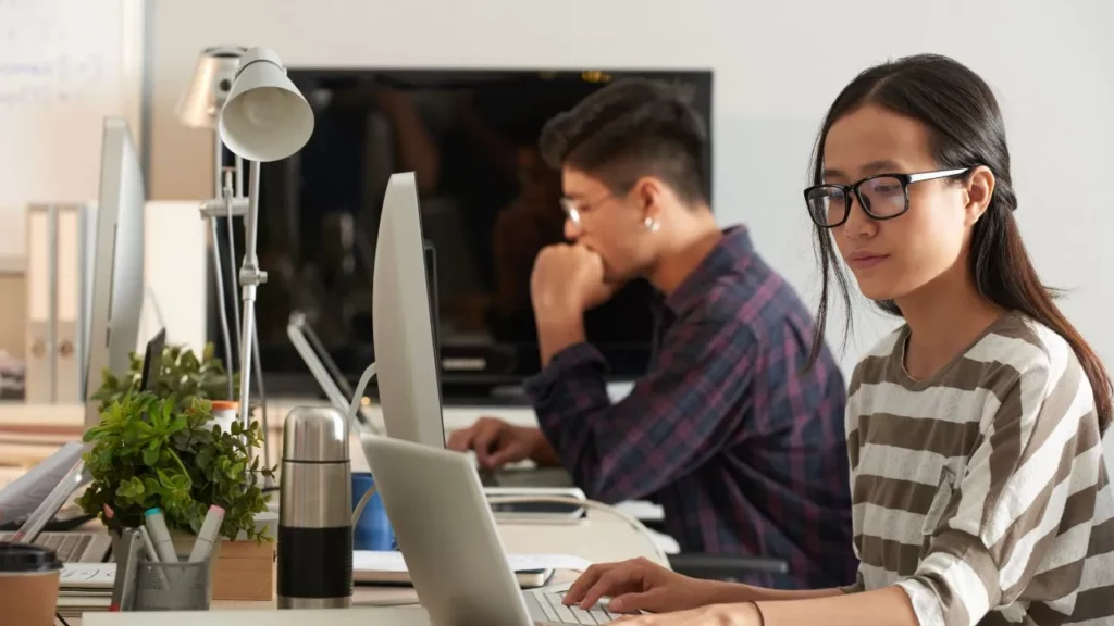 A young woman with glasses focusing on her laptop in a bright, shared office space with a colleague in the background.