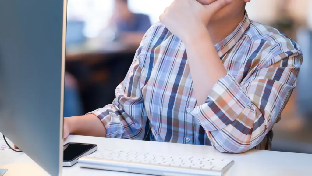 A close-up of a person in a plaid shirt sitting at a desk with their hand to their chin, looking at a computer monitor.