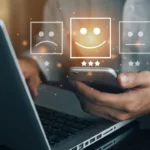 Close-up of a person using a laptop and phone with digital icons of sad, neutral, and happy faces floating above.
