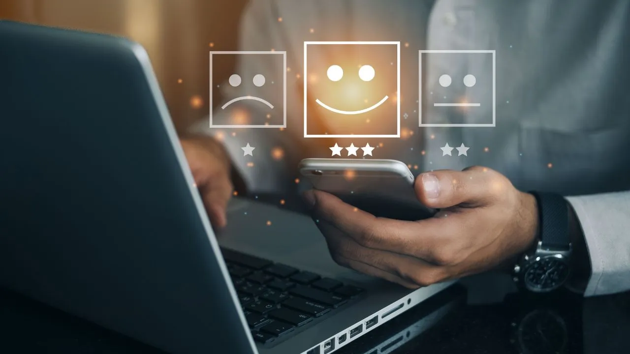 Close-up of a person using a laptop and phone with digital icons of sad, neutral, and happy faces floating above.