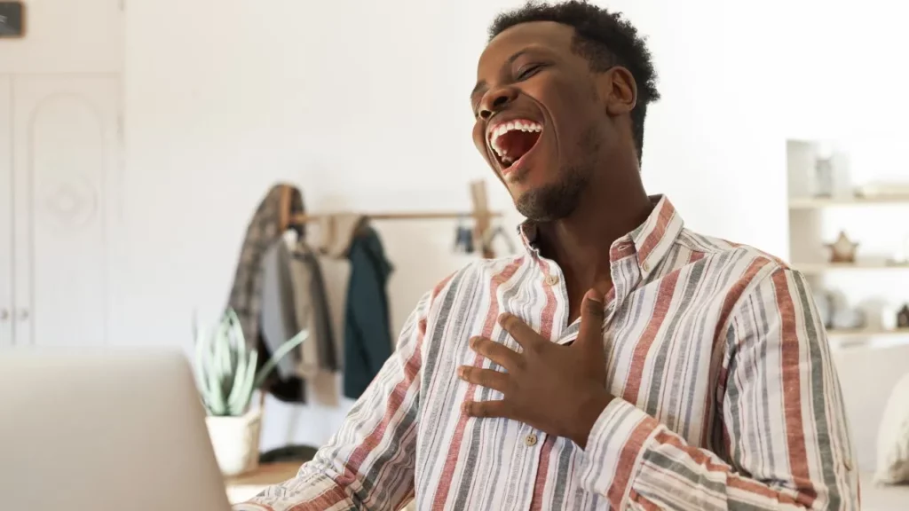 A man in a striped shirt laughing heartily while looking at his laptop screen at home.