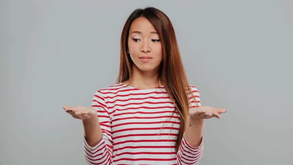 A woman with long brown hair looking down at her empty, upturned palms in a questioning gesture.
