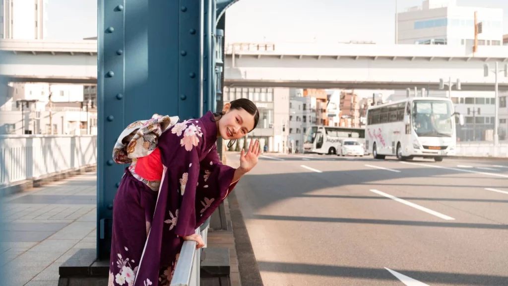 A woman in a dark purple floral kimono waving while leaning against a blue bridge railing.