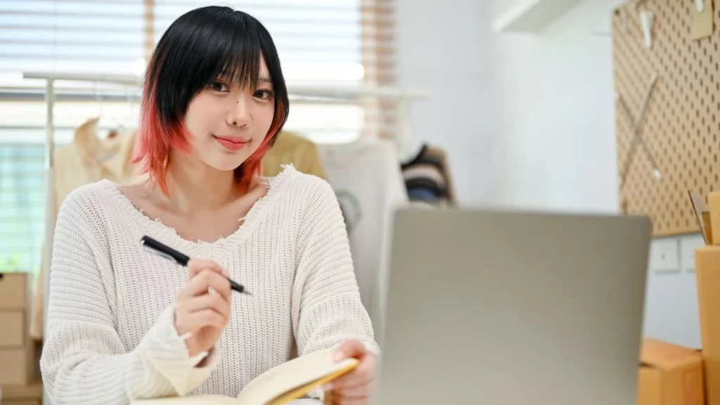 A woman with red-tipped black hair holding a pen and notebook while sitting in front of a laptop.