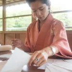 A woman in a pink kimono focused on arranging papers at a wooden table in a traditional Japanese room.