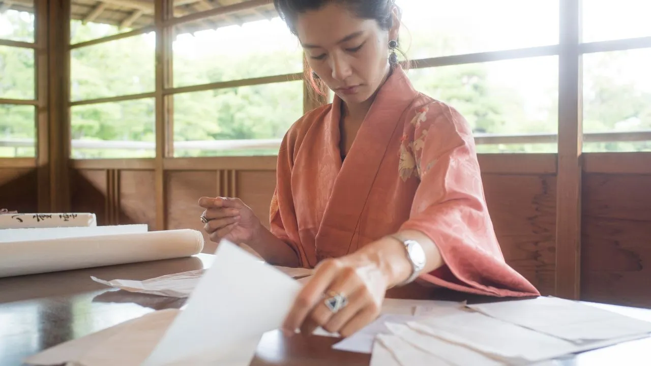 A woman in a pink kimono focused on arranging papers at a wooden table in a traditional Japanese room.