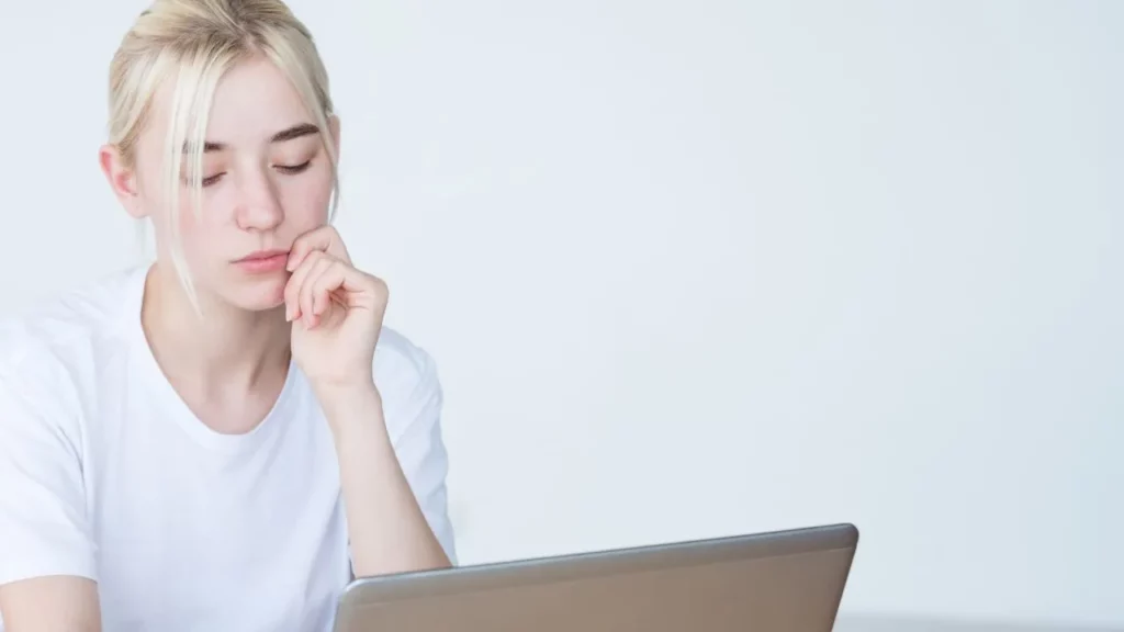 A high-angle photo of a blonde woman looking down thoughtfully at her laptop screen against a plain white background.
